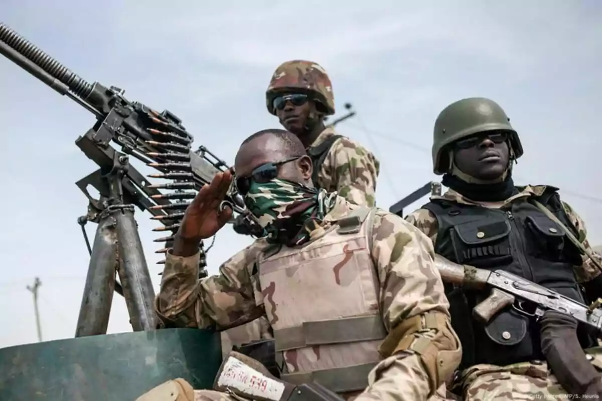 Tres soldados con uniforme de camuflaje y chalecos antibalas están sentados junto a una ametralladora, uno de ellos saluda mientras lleva un pañuelo en el rostro y gafas oscuras