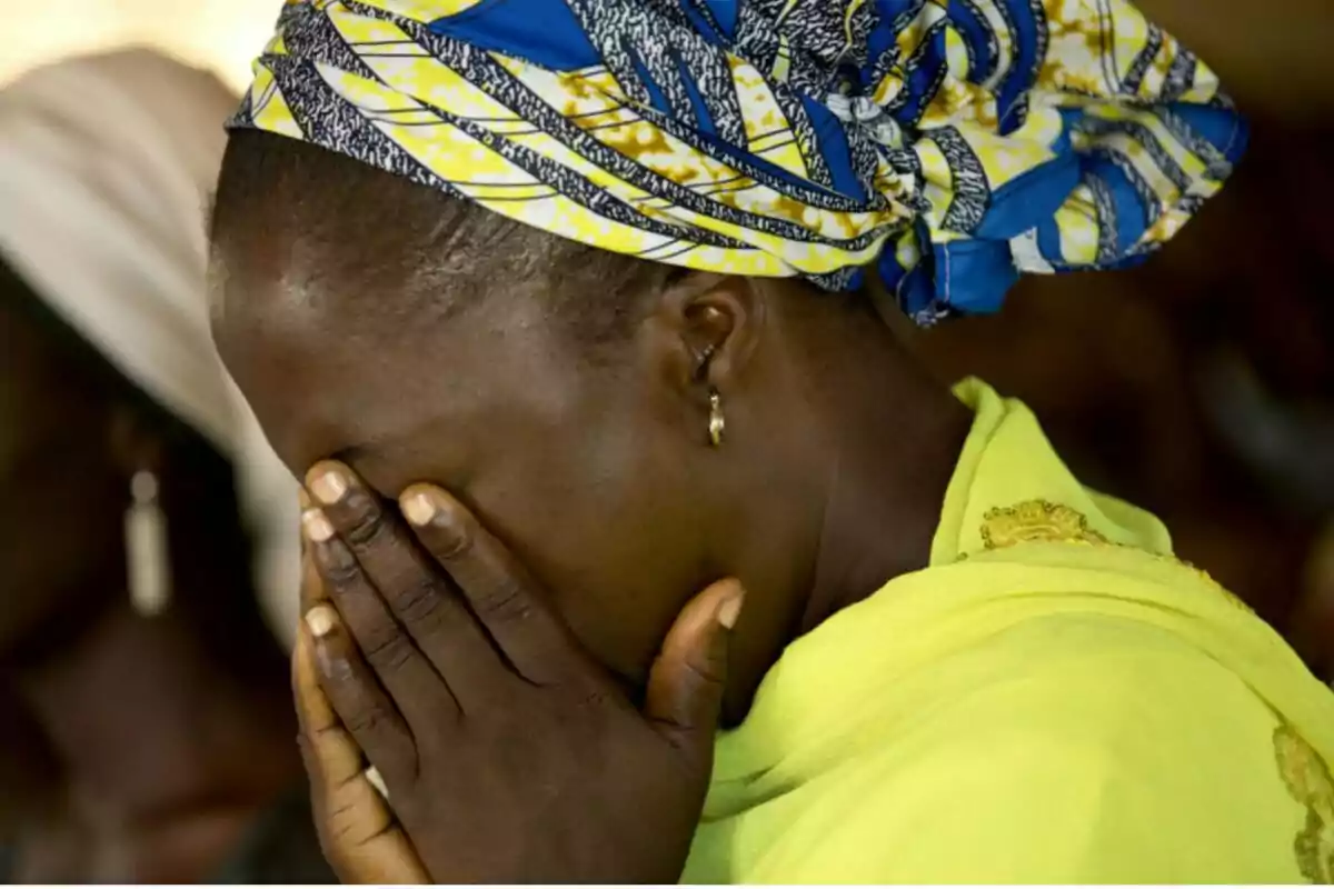 Mujer con pañuelo colorido cubriéndose la cabeza y el rostro con las manos, vestida con ropa amarilla, mostrando una expresión de tristeza o angustia.