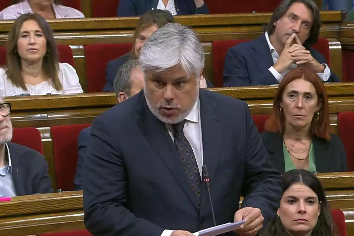 Un home de cabells canosos i barba està parlant en un parlament mentre diverses persones l’observen atentament des dels seus seients.