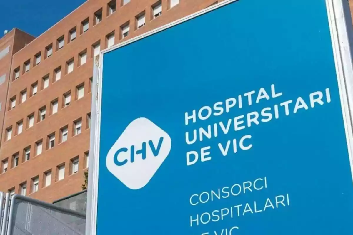 Cartell blau de l’Hospital Universitari de Vic amb un edifici de maó al fons