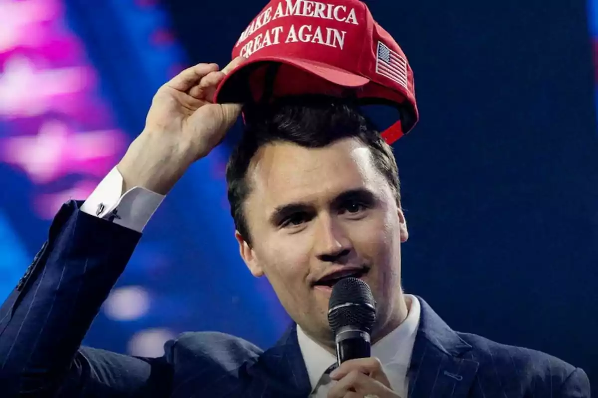 Hombre con micrófono levantando una gorra roja con la frase Make America Great Again en un escenario
