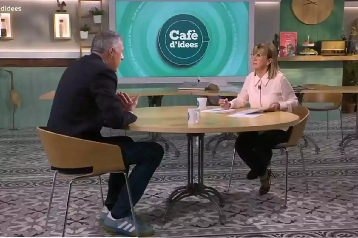 Dos personas sentadas en una mesa redonda conversan en un set de televisión decorado con tonos verdes y un gran monitor al fondo que muestra el logo de Café d’idees Dos personas sentadas en una mesa redonda conversan en un set de televisión decorado con tonos verdes y un gran monitor al fondo que muestra el logo de Café d’idees