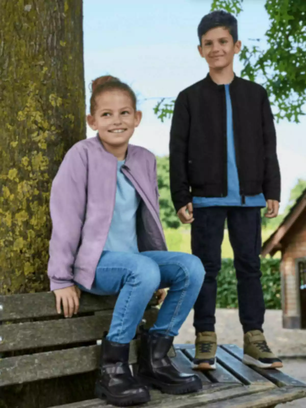 Dos nens amb jaquetes i texans posen a l’aire lliure al costat d’un arbre i un banc de fusta.