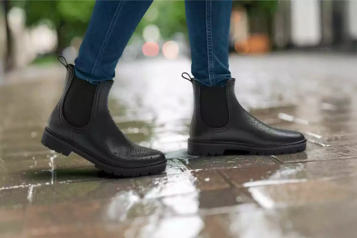 Persona usando botas negras impermeables y pantalón de mezclilla caminando sobre una acera mojada Persona usando botas negras impermeables y pantalón de mezclilla caminando sobre una acera mojada