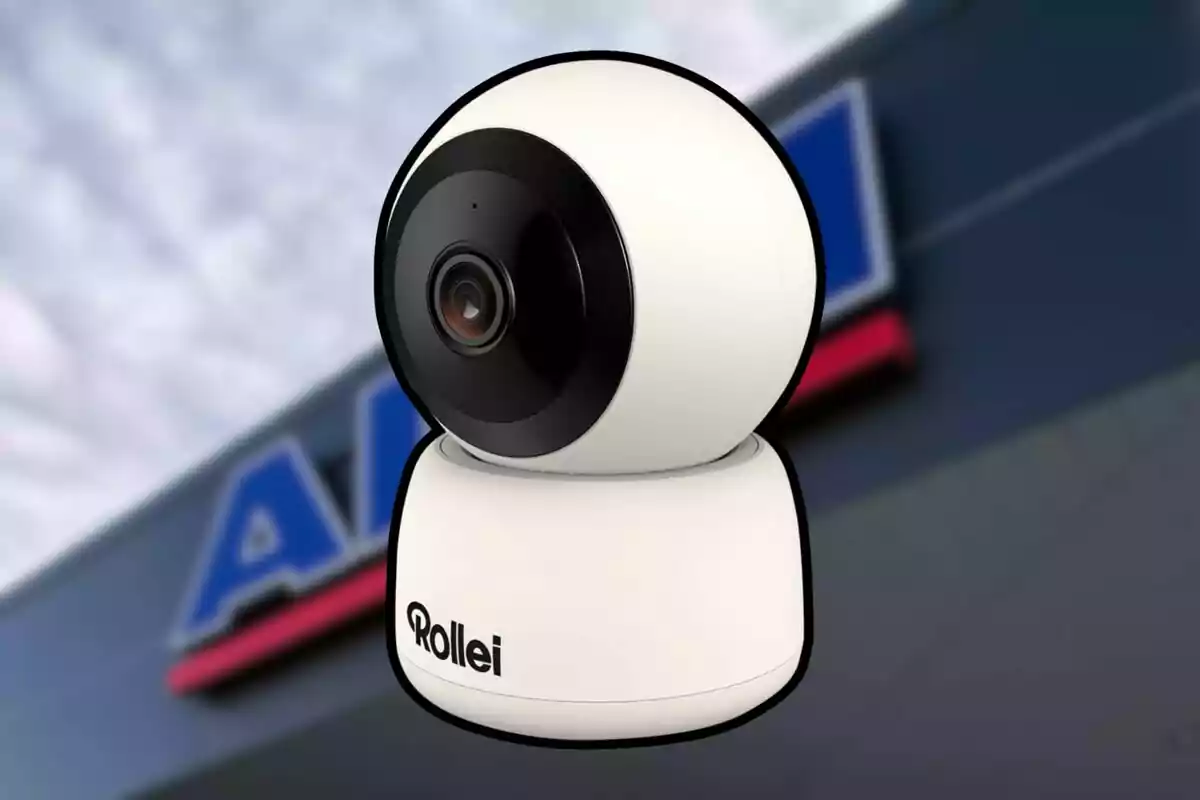 Cámara de seguridad blanca marca Rollei con fondo desenfocado de un letrero azul y rojo.