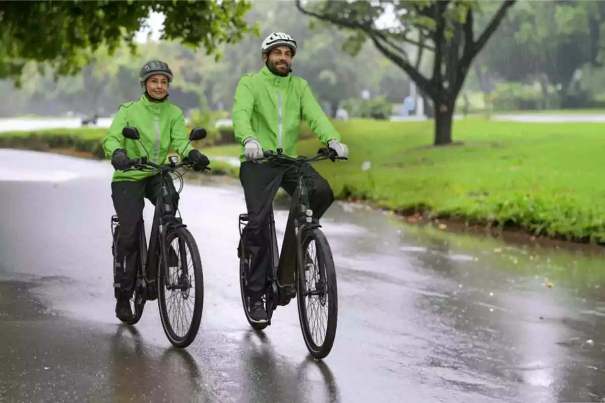 Dos personas con chaquetas verdes y cascos montan bicicletas en un camino mojado rodeado de árboles y césped Dos personas con chaquetas verdes y cascos montan bicicletas en un camino mojado rodeado de árboles y césped
