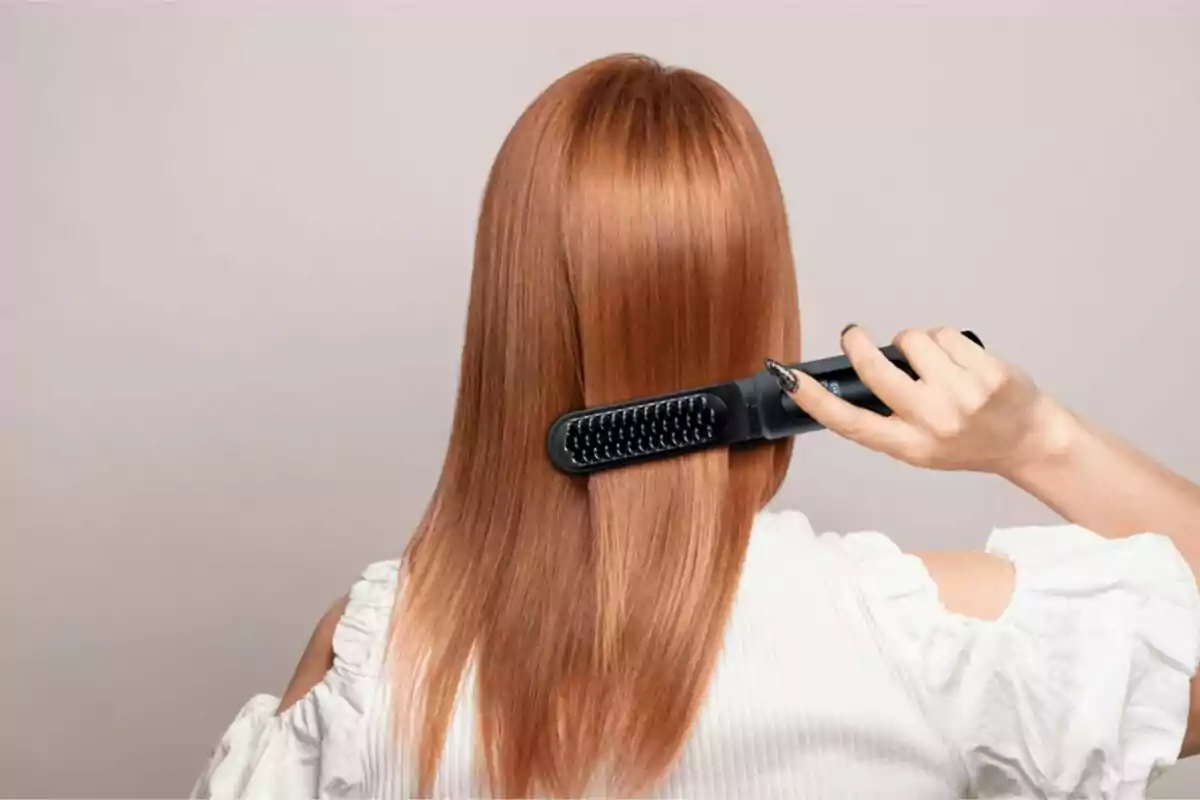 Mujer de espaldas alisando su cabello lacio y pelirrojo con un cepillo eléctrico
