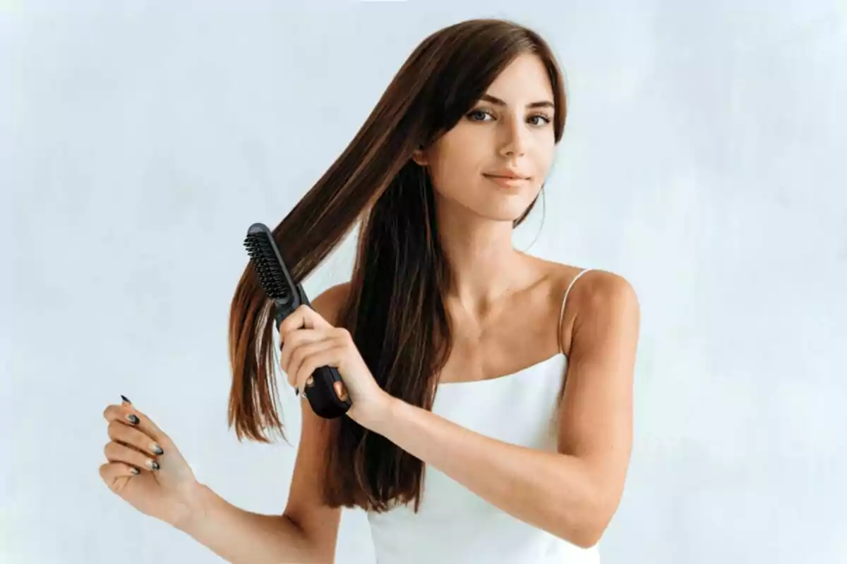 Mujer joven con cabello largo y liso usando un cepillo eléctrico para peinarse mientras sonríe suavemente