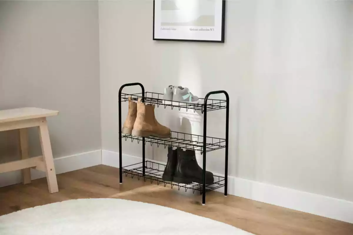 Zapatero de metal negro con tres niveles que sostiene botas marrones, botas negras y unos tenis grises en una habitación minimalista con piso de madera