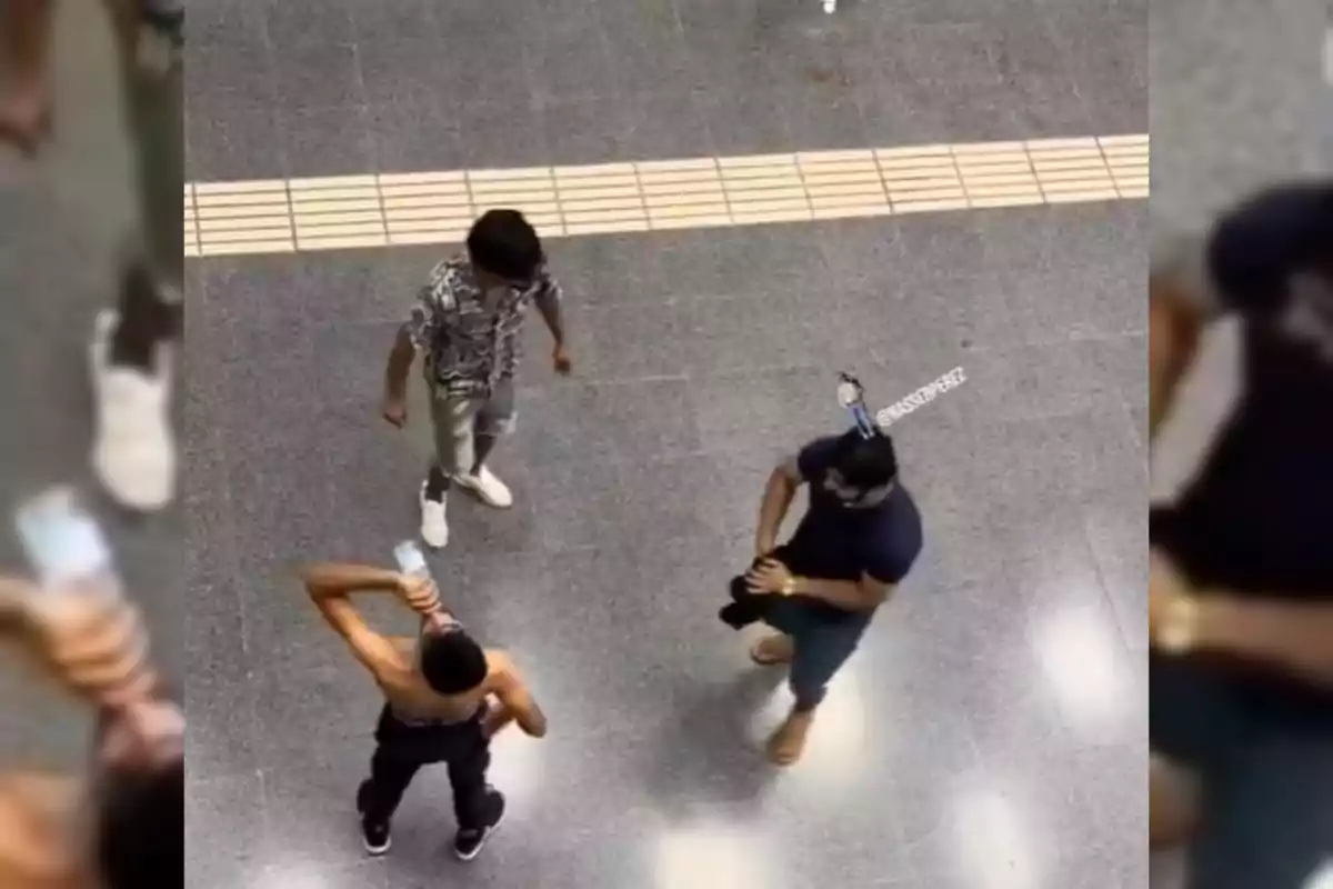 Tres personas están de pie en el suelo gris de una estación, una de ellas sin camiseta bebiendo de una botella, mientras las otras dos observan