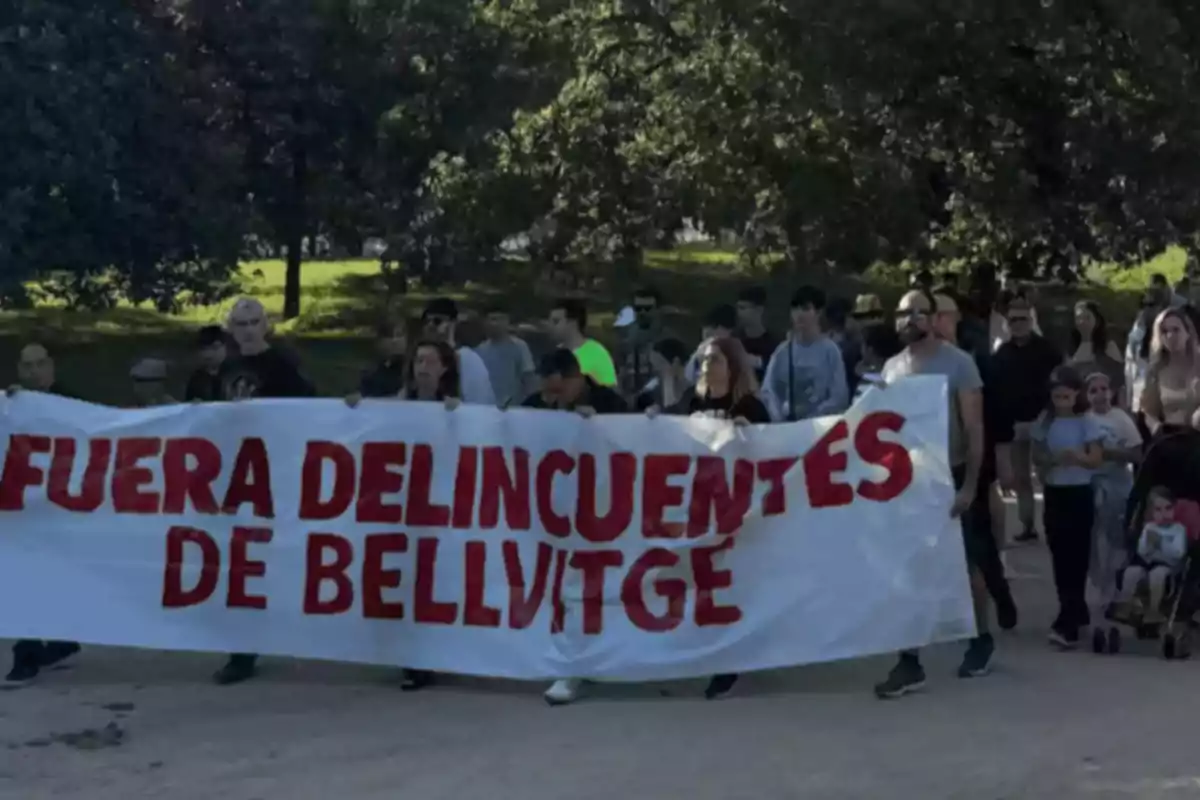 Un grup de persones marxa per un parc sostenint una pancarta blanca amb lletres vermelles que diu "Fora delinqüents de Bellvitge"
