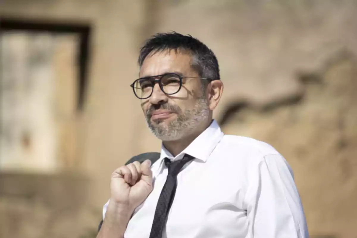 Hombre de mediana edad con barba y gafas vistiendo camisa blanca y corbata negra al aire libre con expresión pensativa