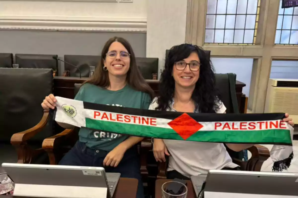 Dos mujeres sonríen sentadas en una sala mientras sostienen una bufanda con los colores de la bandera de Palestina y la palabra Palestine escrita en ambos extremos