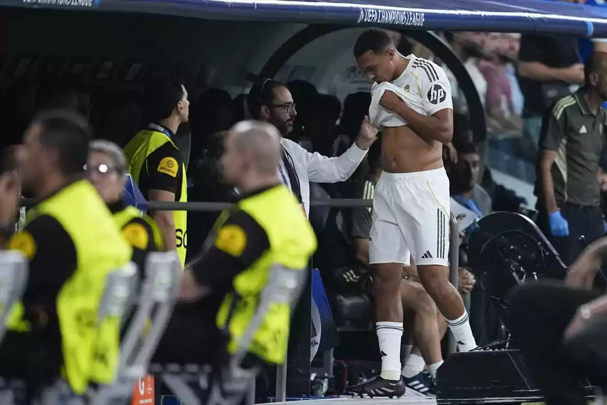Un futbolista del Reial Madrid és revisat per un metge a la zona de banqueta mentre s’aixeca la samarreta mostrant l’abdomen durant un partit, amb personal de seguretat i altres jugadors al voltant.
