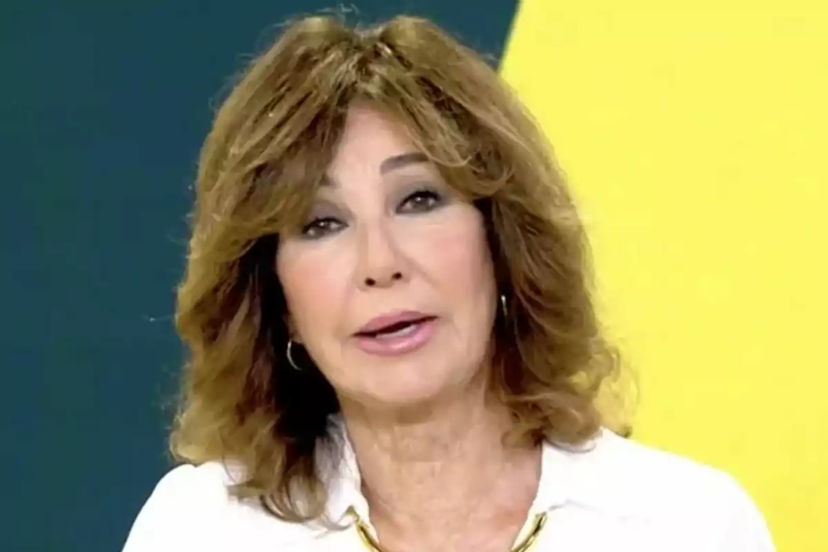 Ana Rosa Quintana de cabello castaño claro y ondulado con camisa blanca y collar dorado sobre un fondo amarillo y azul oscuro en El Programa de Ana Rosa.