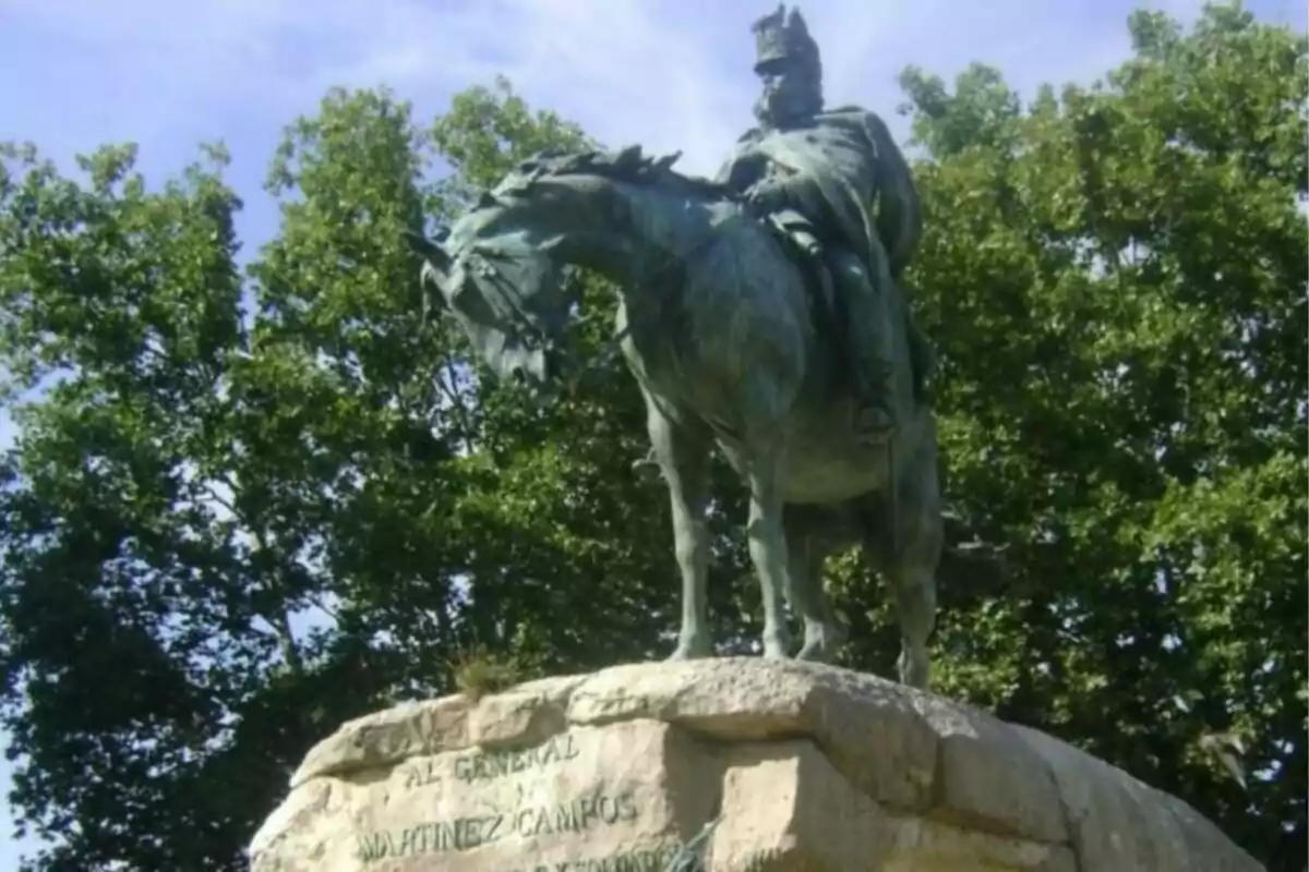 Estàtua eqüestre d’un home amb uniforme militar sobre un pedestal de pedra envoltat d’arbres i cel blau Estàtua eqüestre d’un home amb uniforme militar sobre un pedestal de pedra envoltat d’arbres i cel blau