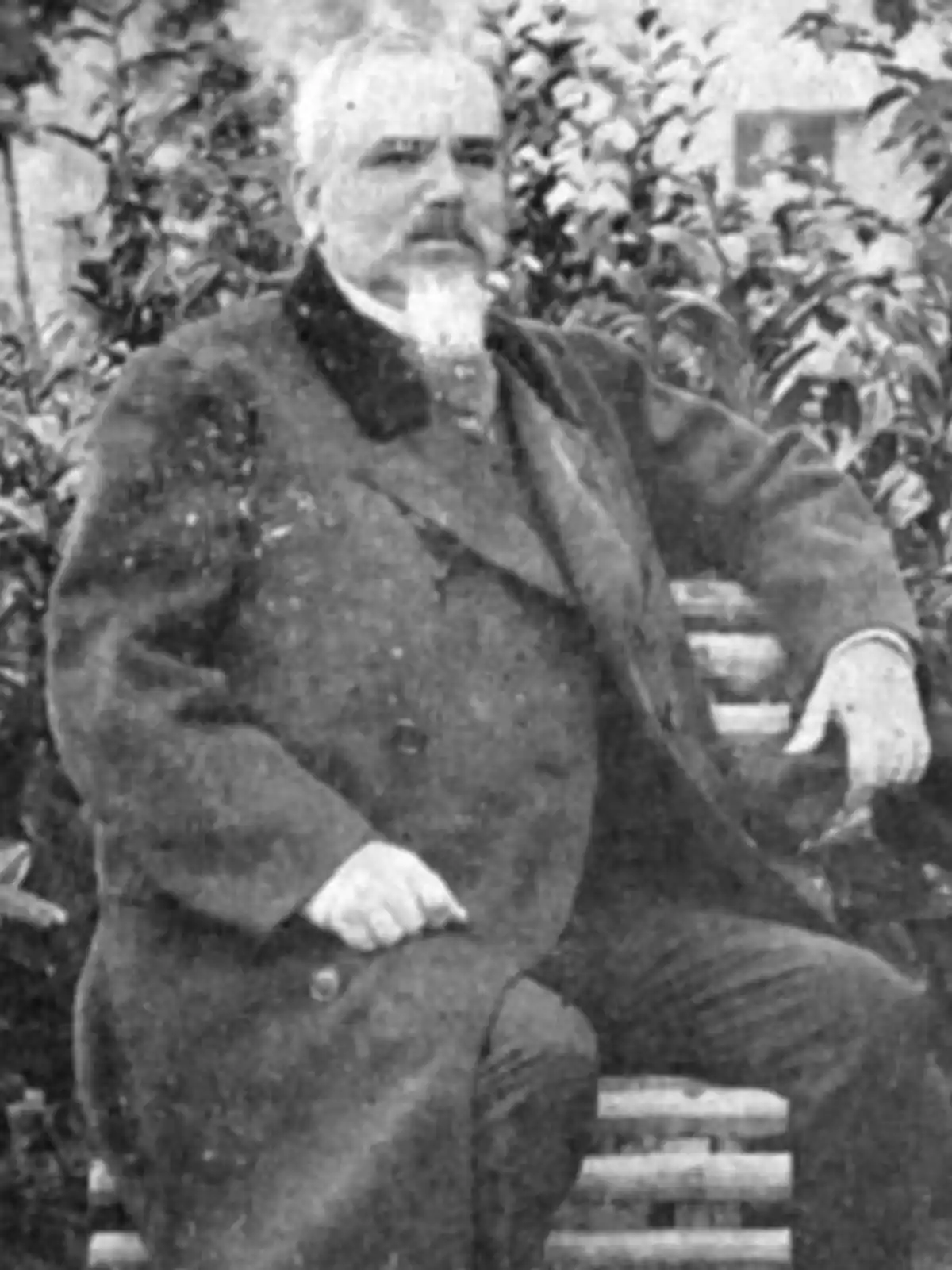 Home gran amb barba assegut en un banc de fusta, vestit amb un abric fosc en un entorn a l’aire lliure amb vegetació al fons. Home gran amb barba assegut en un banc de fusta, vestit amb un abric fosc en un entorn a l’aire lliure amb vegetació al fons.