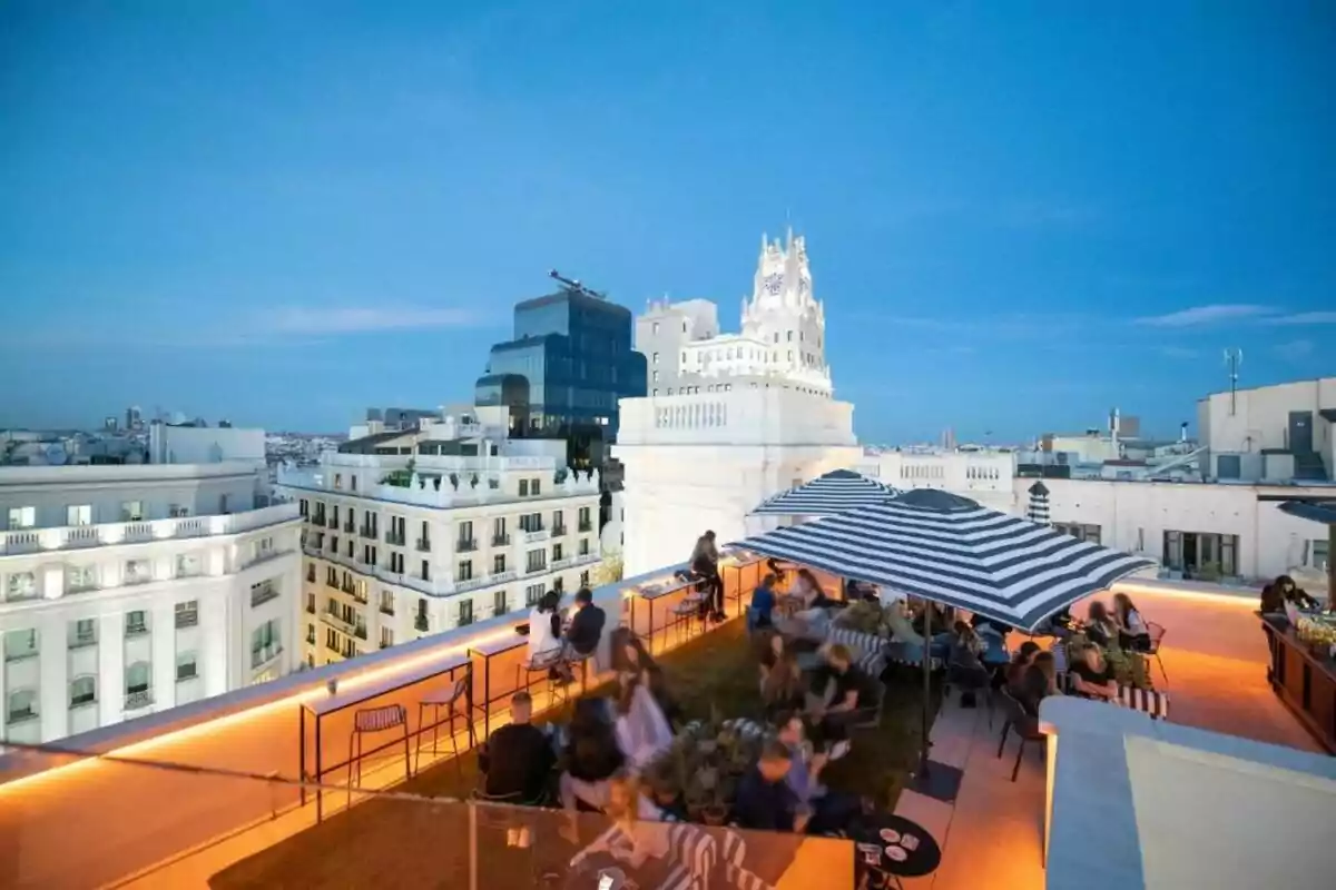 Persones gaudint en una terrassa a la teulada d’un edifici amb para-sols de ratlles i vistes a la ciutat al capvespre Persones gaudint en una terrassa a la teulada d’un edifici amb para-sols de ratlles i vistes a la ciutat al capvespre