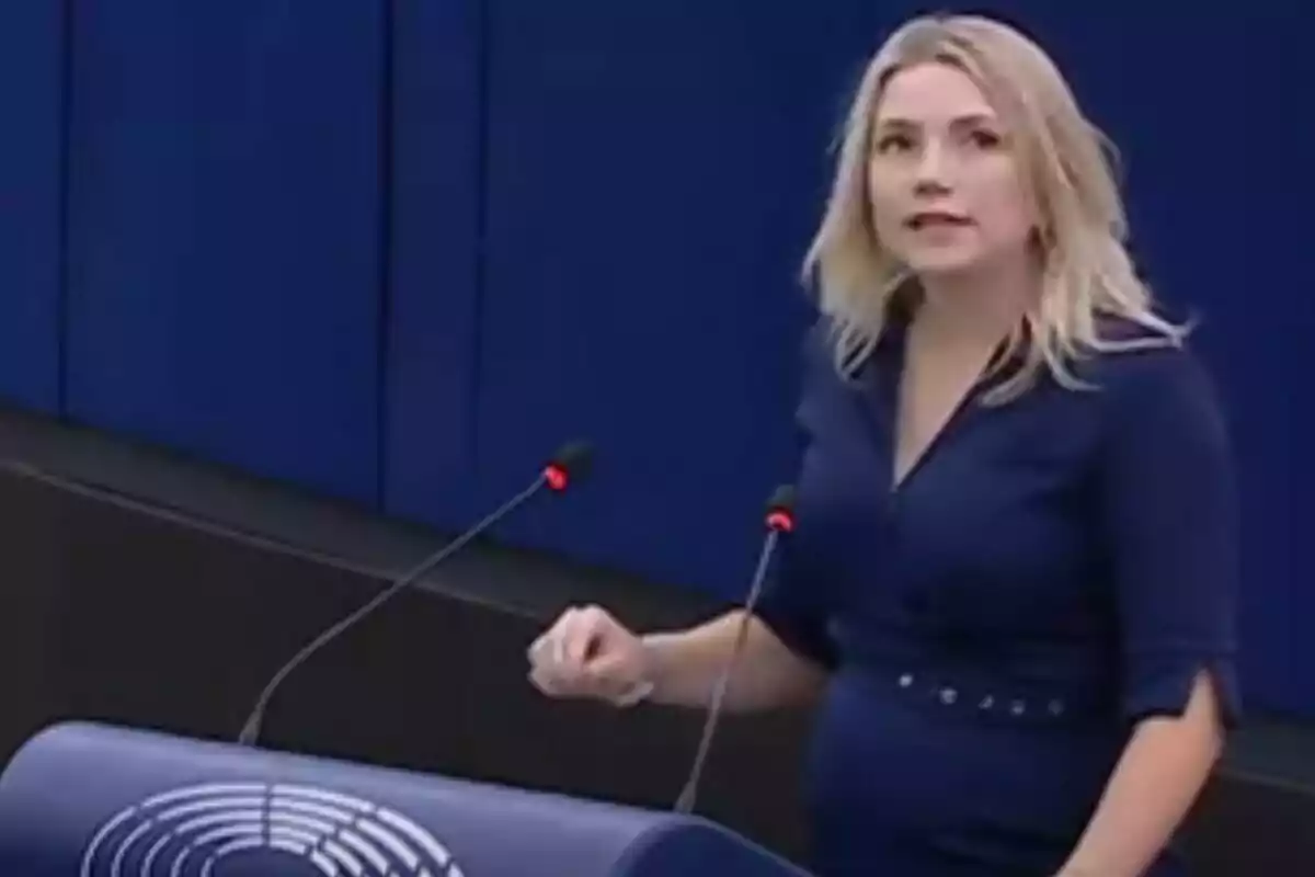 Mujer rubia hablando frente a un podio con micrófonos en un entorno institucional de fondo azul