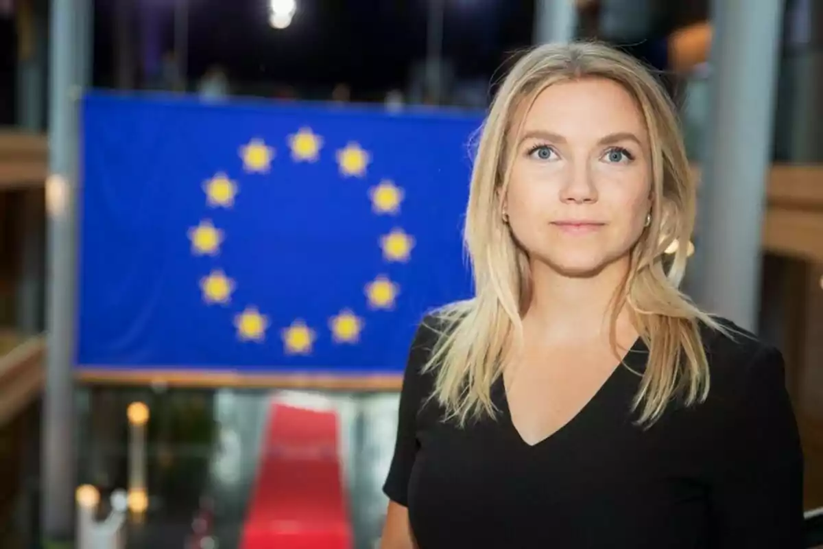 Mujer rubia con camiseta negra posando frente a una bandera de la Unión Europea en un edificio moderno