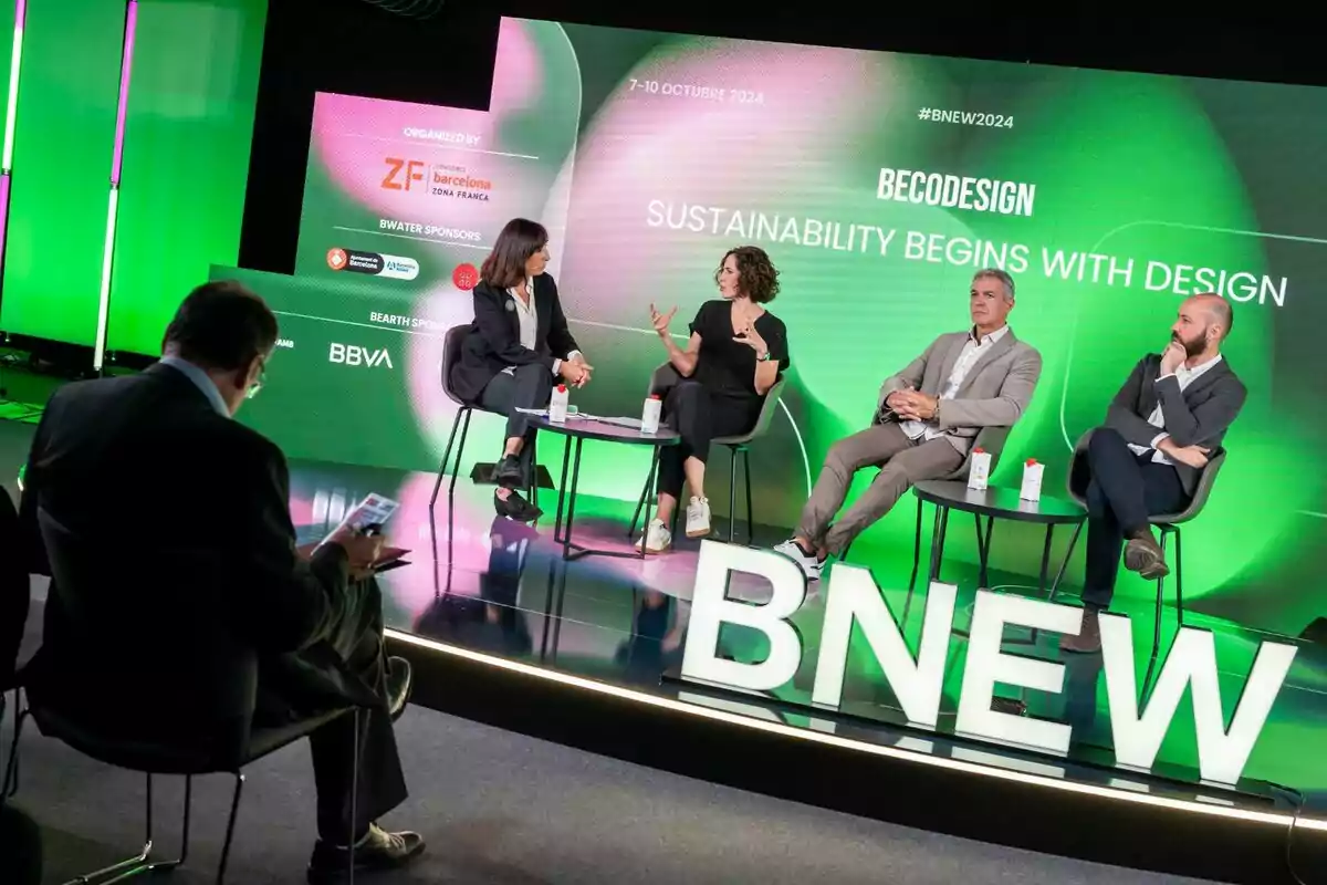Cuatro personas participan en un panel de discusión sobre sostenibilidad y diseño en un escenario con fondo verde y letras grandes de BNEW mientras un hombre en primer plano observa y toma notas.