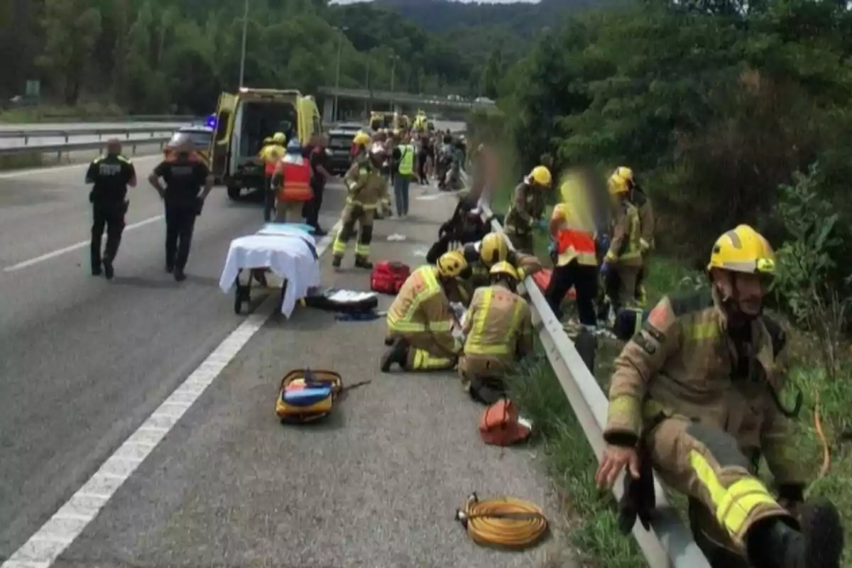 Diversos bombers i personal d’emergència atenen un accident en una carretera al costat d’ambulàncies i patrulles, mentre alguns rescatadors ajuden persones ferides a prop de la bardissa.