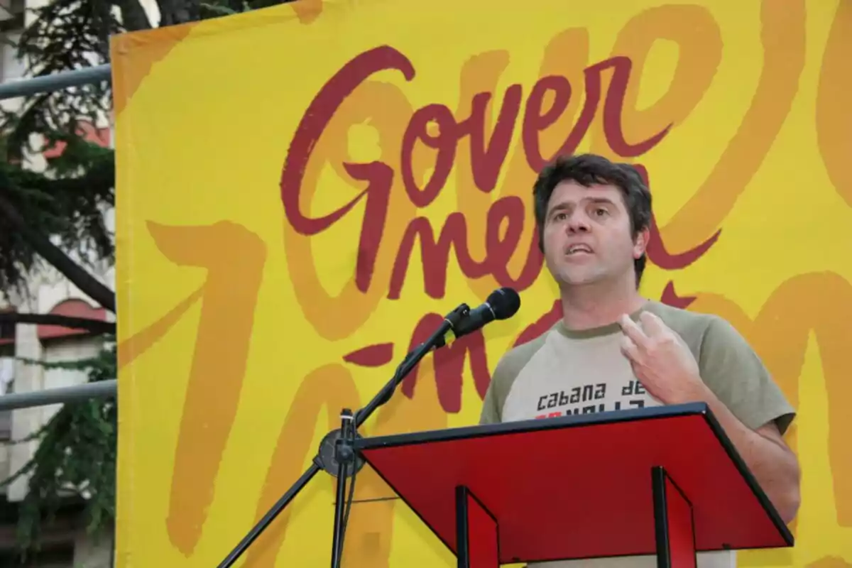 Home parlant en un faristol vermell davant d’un fons groc amb lletres grans i colorides