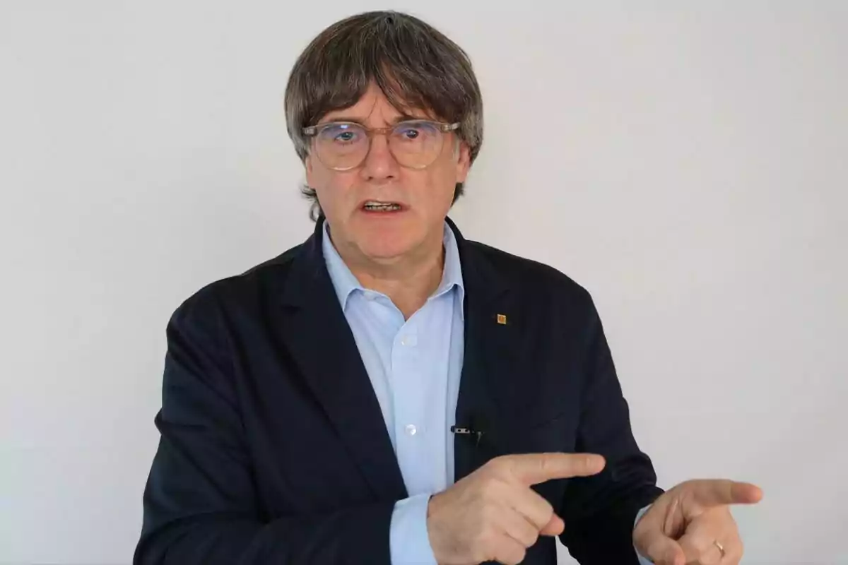 Hombre de cabello canoso y gafas con chaqueta azul y camisa clara señalando con ambas manos frente a un fondo blanco