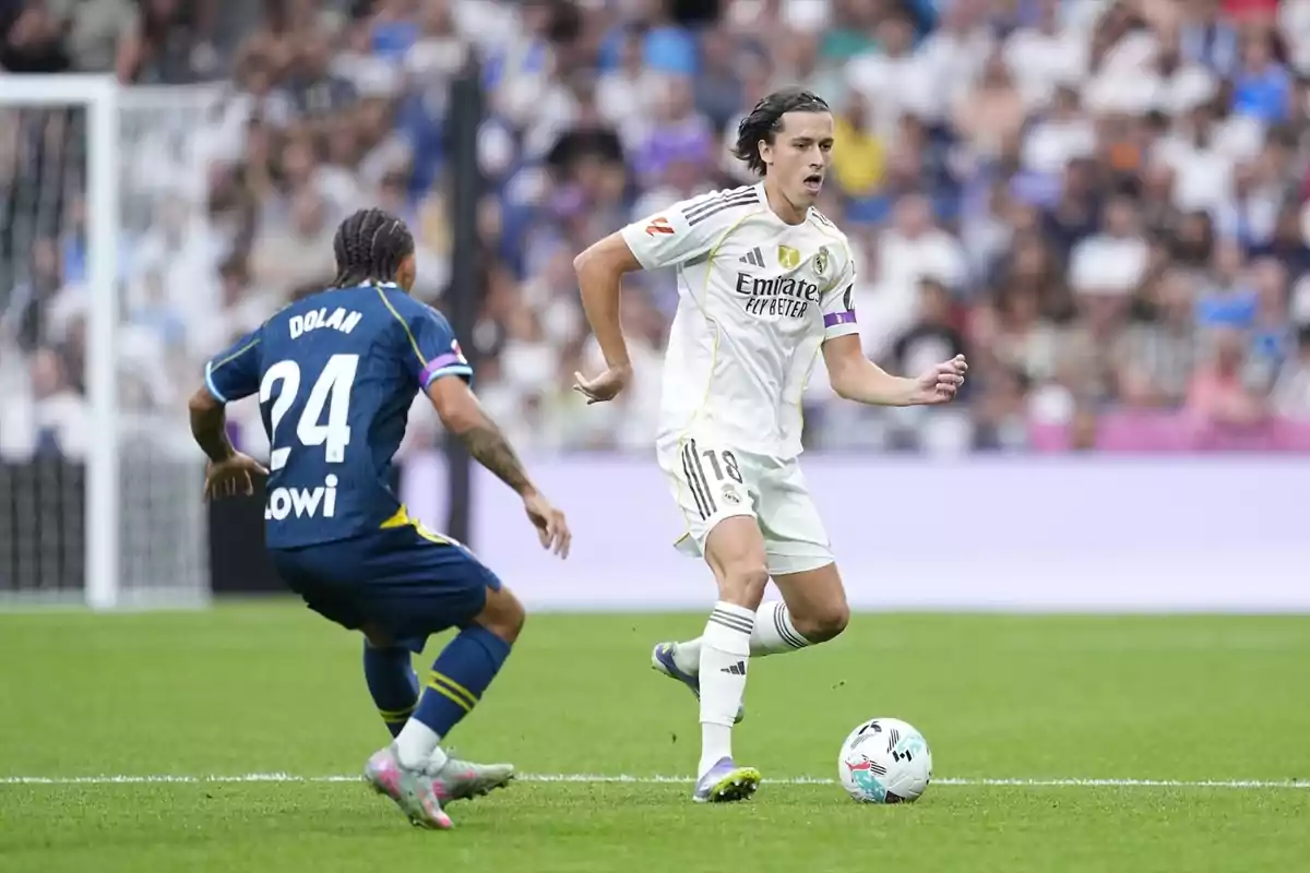Dos futbolistes disputen la pilota durant un partit en un estadi ple d’espectadors, un amb uniforme blanc i l’altre amb uniforme blau.