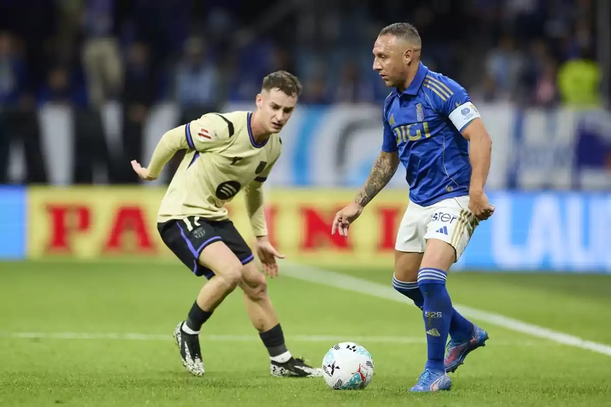 Dos futbolistes disputen la pilota durant un partit en un estadi, un amb uniforme blau i l’altre amb uniforme crema.