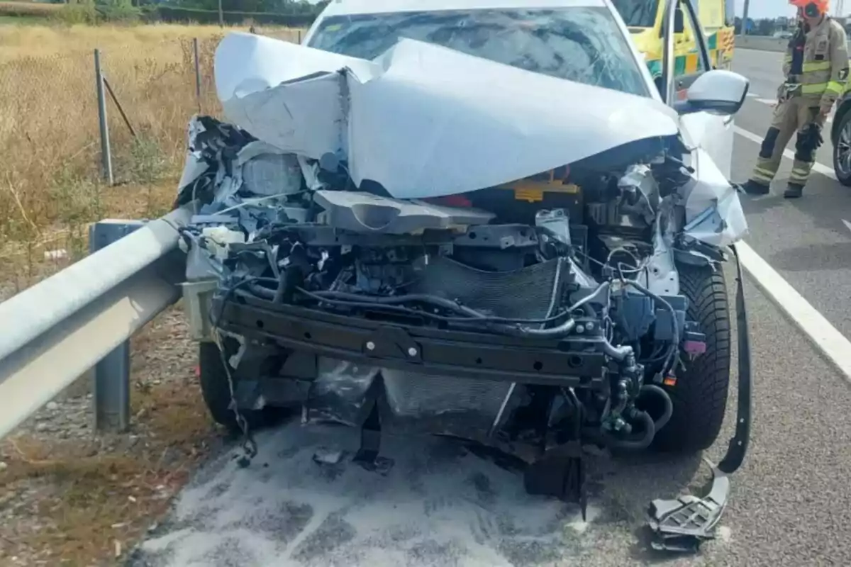 Un cotxe blanc amb la part davantera completament destrossada després d’un accident a l’A-2, amb un bomber i una ambulància al fons.