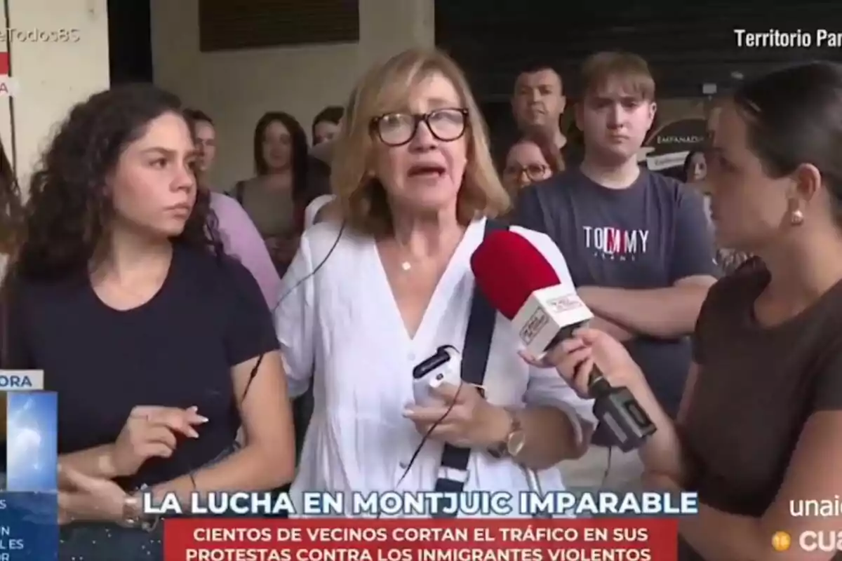 Una reportera entrevista una dona gran amb ulleres i samarreta blanca mentre diverses persones observen al fons i a la part inferior de la imatge apareix un titular de notícies sobre protestes a Montjuïc.