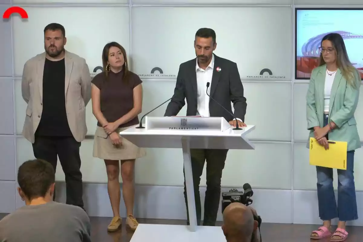 Quatre persones dretes en una sala de premsa del Parlament de Catalunya mentre una d’elles parla en un faristol i dues altres persones fan fotos o graven des del públic.