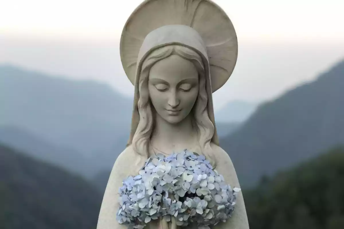 Escultura d’una figura religiosa femenina sostenint un ram de flors blaves amb muntanyes al fons