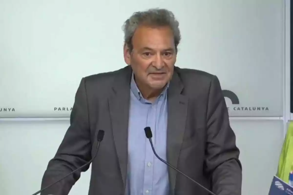 Hombre de cabello canoso y saco gris hablando frente a dos micrófonos en una sala de conferencias