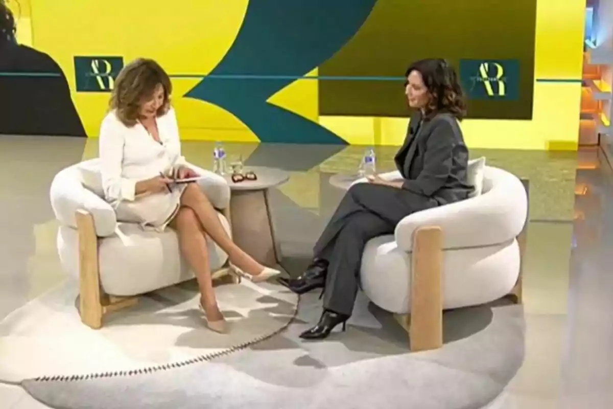 Dos mujeres sentadas en sillones blancos conversan en un set de televisión con fondo amarillo y detalles en verde, una de ellas viste de blanco y la otra de gris oscuro, entre ellas hay una mesa pequeña con botellas de agua y gafas; son Isabel Díaz Ayuso y Ana Rosa Quintana. Dos mujeres sentadas en sillones blancos conversan en un set de televisión con fondo amarillo y detalles en verde, una de ellas viste de blanco y la otra de gris oscuro, entre ellas hay una mesa pequeña con botellas de agua y gafas; son Isabel Díaz Ayuso y Ana Rosa Quintana.