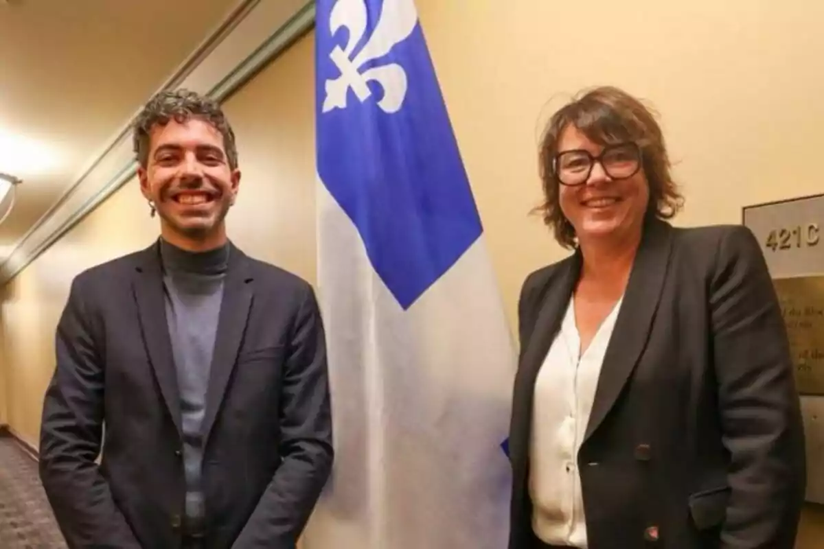 Dos personas sonrientes posan junto a una bandera azul y blanca con un símbolo de flor de lis en un pasillo iluminado