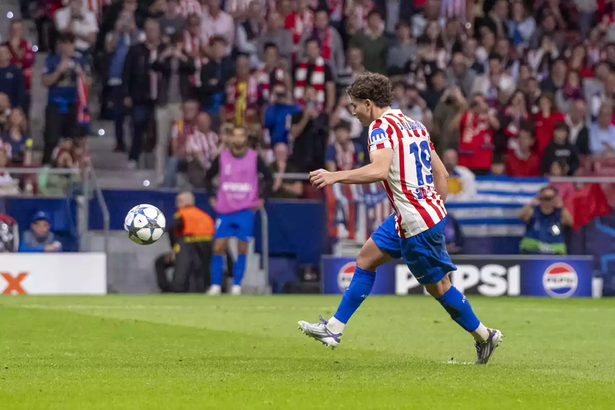 Atlético de Madrid player in a red, white, and blue uniform kicks a ball during a match in a stadium full of fans.