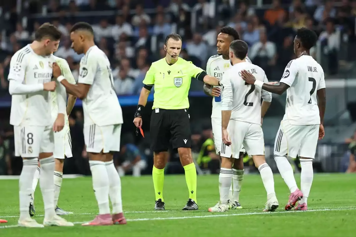 Un àrbitre de futbol sosté una targeta vermella mentre diversos jugadors del Reial Madrid l’envolten al camp durant un partit.