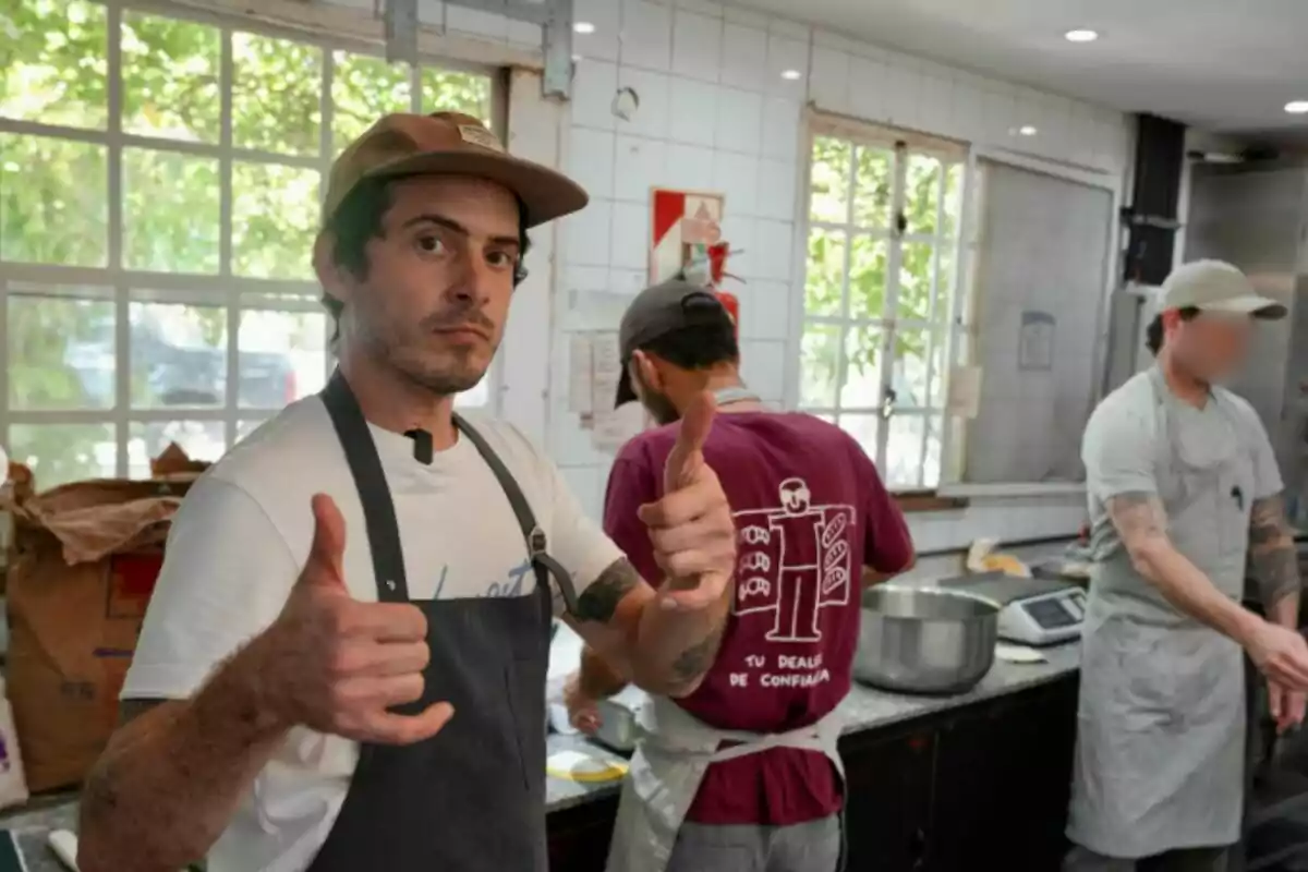 Home amb gorra i davantal fent el gest de polze amunt en una cuina al costat de dues persones més treballant