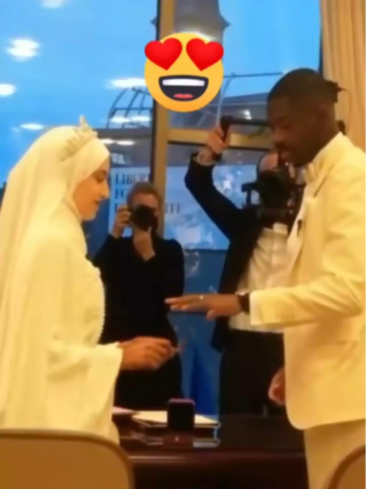 Ousmane Dembéle i Rima Edbouche vestits de blanc intercanviant anells en una cerimònia de casament amb fotògrafs al fons i un emoji d’ulls de cor sobre ells Ousmane Dembéle i Rima Edbouche vestits de blanc intercanviant anells en una cerimònia de casament amb fotògrafs al fons i un emoji d’ulls de cor sobre ells