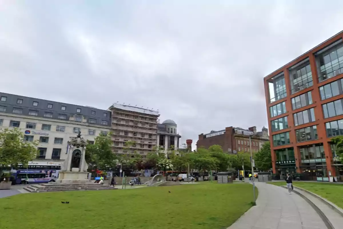 Plaça amb gespa envoltada d’edificis moderns i antics, amb una estàtua al centre i persones passejant pels camins a Manchester.