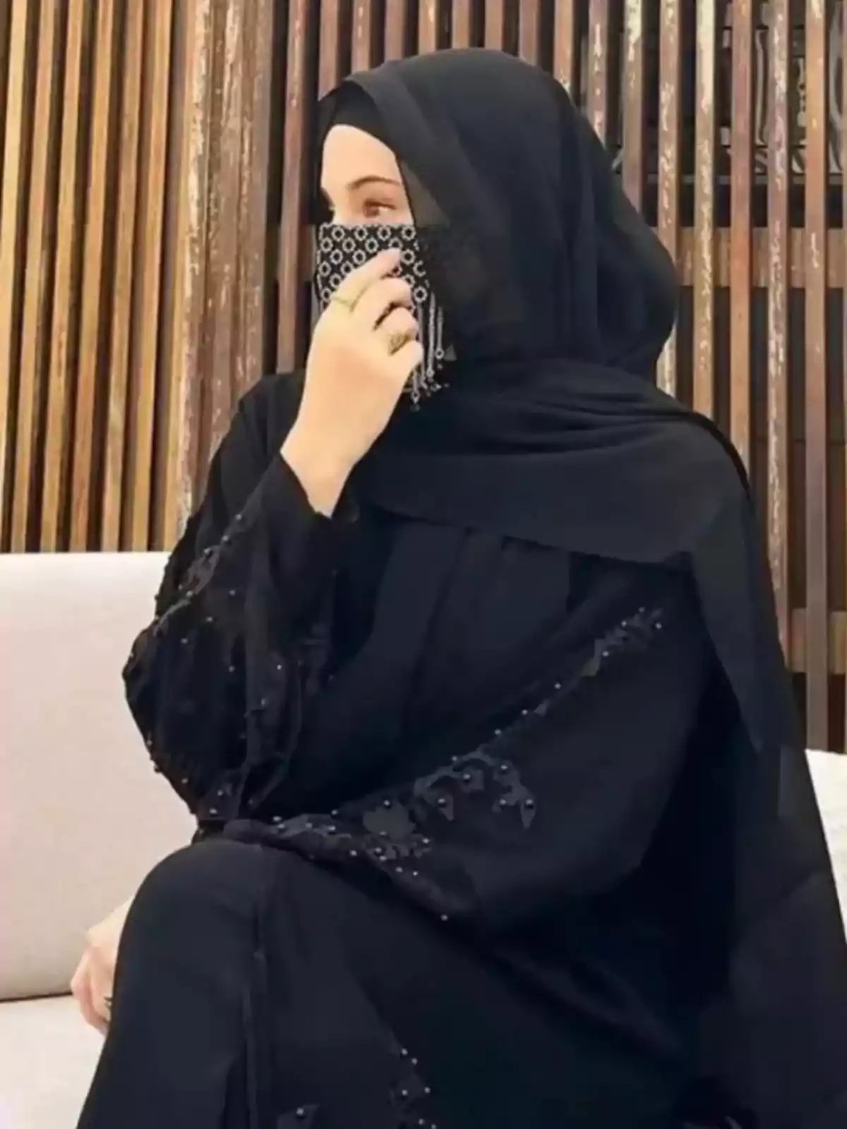 Rima Edbouche asseguda amb un hijab negre i mascareta decorada, mirant cap a un costat sobre un fons de llistons de fusta.