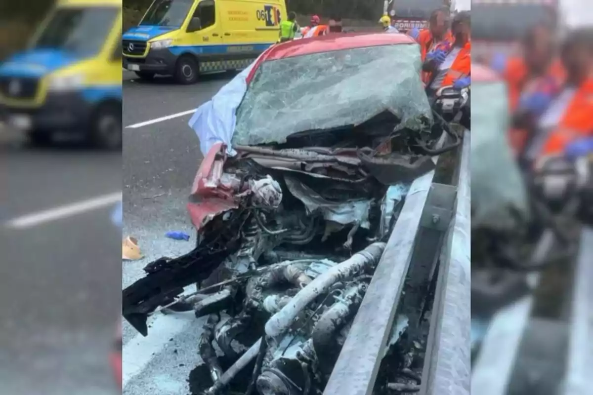 Coche rojo gravemente accidentado en una carretera con el frente completamente destruido y personal de emergencia atendiendo la situación