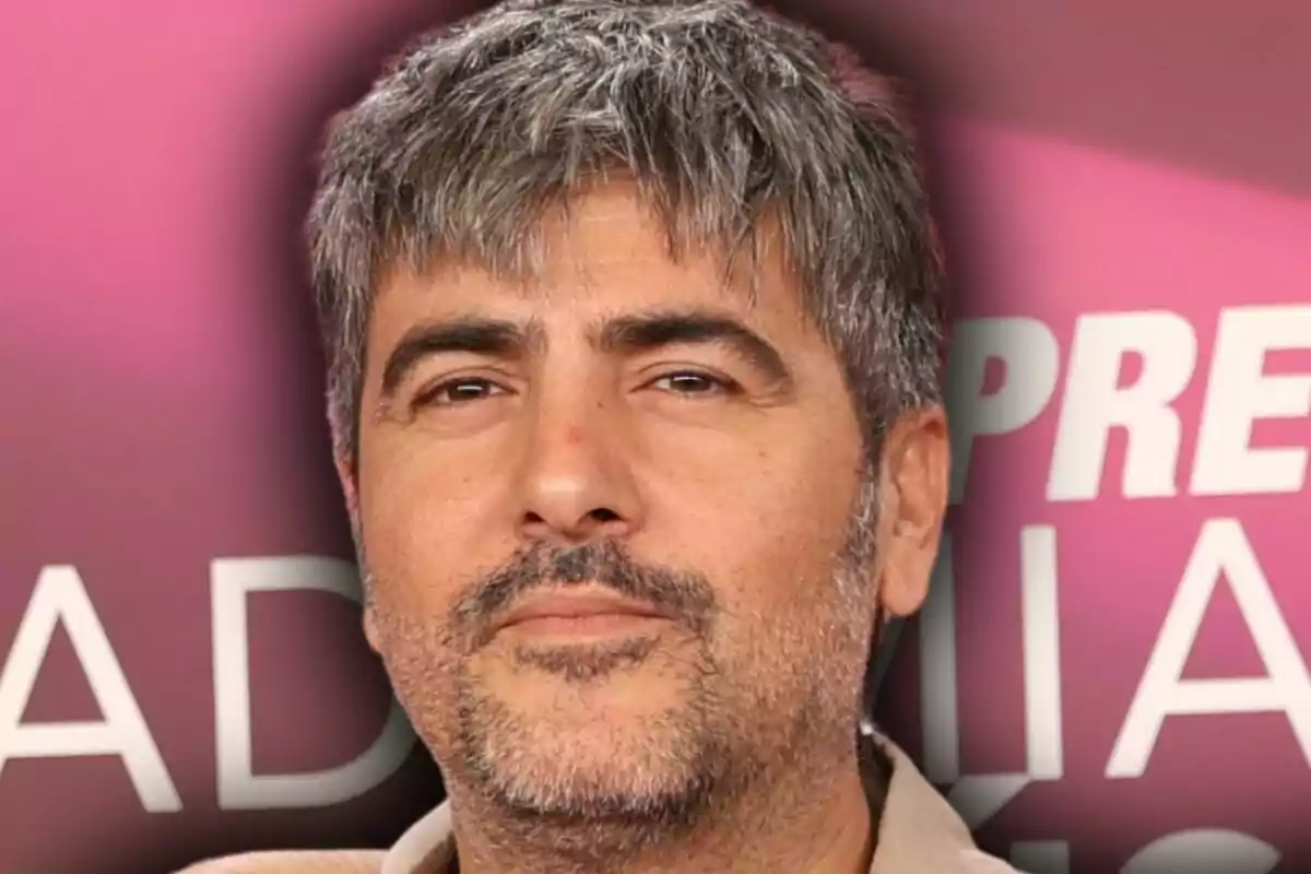 Home de cabells canosos i barba curta posant davant d’un fons rosa amb lletres blanques desenfocades