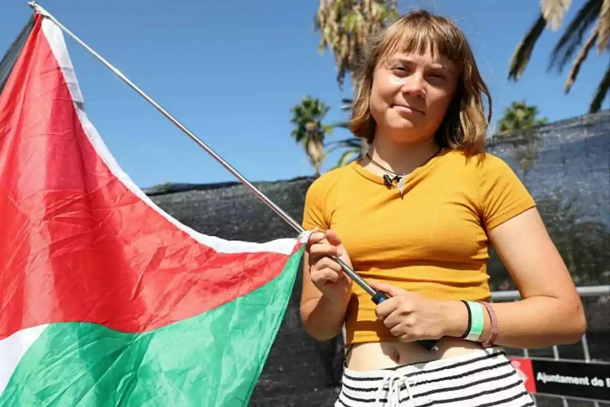 Joven con camiseta amarilla sostiene una bandera de Palestina al aire libre con palmeras al fondo Joven con camiseta amarilla sostiene una bandera de Palestina al aire libre con palmeras al fondo