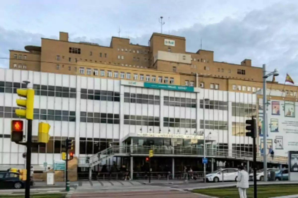 Façana de l'Hospital Universitari Miguel Servet de Saragossa, on s'observen diverses plantes, persones i cotxes a l'entrada i un semàfor en vermell en primer pla.