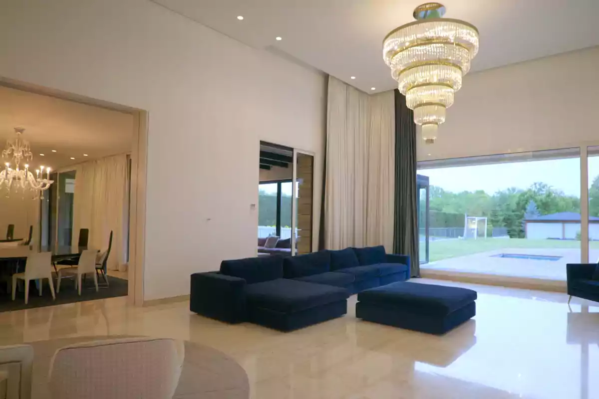 Sala moderna amb sofà blau, làmpada de cristall gran, finestral amb vista al jardí i menjador al fons Sala moderna amb sofà blau, làmpada de cristall gran, finestral amb vista al jardí i menjador al fons