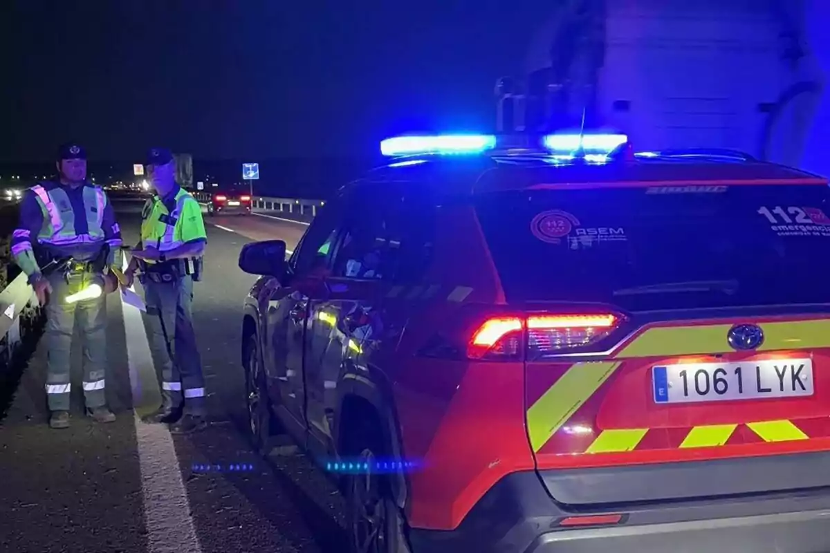 Dos agentes de tráfico conversan junto a un vehículo de emergencias con luces encendidas en una carretera durante la noche