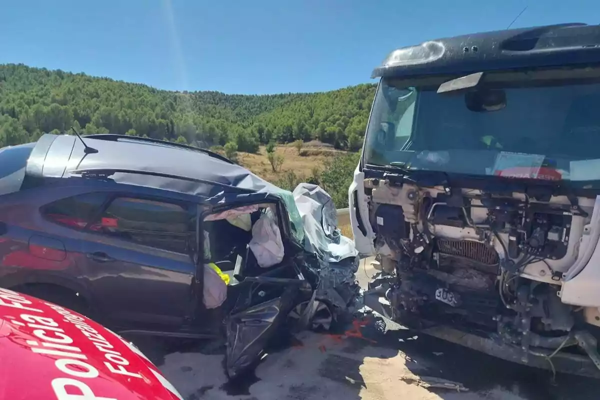 Accidente vial entre un coche negro y un camión blanco en una carretera rodeada de árboles, con daños severos en ambos vehículos y presencia policial